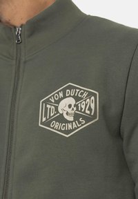 Veste zippée vert olive en tissu sweat, dotée d'un grand logo crâne crème dans un badge hexagonal sur la poitrine.