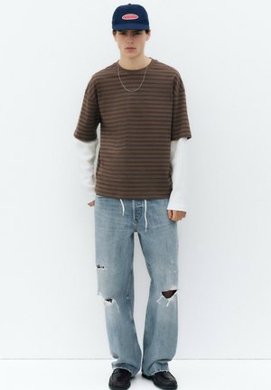 Ung mand iført en brunstribet t-shirt over en lang hvid skjorte, revnede lyseblå jeans, sorte sko og en marineblå baseballkasket.