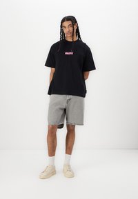 Black cotton t-shirt with a pink graphic print, paired with light gray denim shorts. Model wears cream sneakers with white socks.