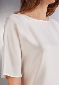 Blusa de satén en crema con mangas cortas y un escote redondeado. Textura suave con un brillo sutil, con líneas simples y limpias.