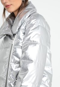 Srebrna kurtka puchowa o błyszczącym wykończeniu, z wysokim kołnierzem, zamkiem błyskawicznym i poziomym przeszyciem dla tekstury.