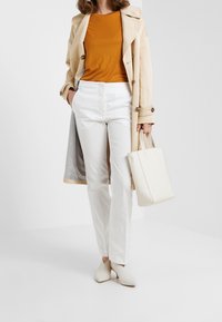 Beige trenčkot přes oranžový top, bílé kalhoty a bílé boty. Oblečení zahrnuje minimalistickou bílou kabelku. Hladké textury, přiléhavý střih.