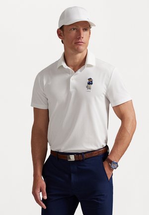 Man wearing white baseball cap, white polo shirt with embroidered bear, navy trousers, brown belt, and wristwatch, posing with one hand in pocket.