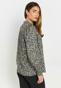 Långärmad blus i svart och krämfärgat blommönster. Har en hög krage och gathered ärmar för en avslappnad passform. Mjuk tygtextur.