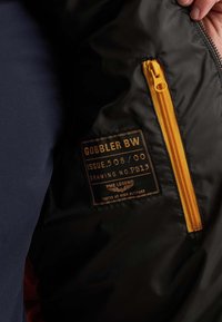 Ręka trzyma otwartą czarną kurtkę, widać pomarańczowy zamek błyskawiczny i metkę z napisem "Gobbler BW, Issue 508/00, PME Legend, Przetestowane na dużej wysokości."