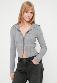 Sivota skrajšana jakna z zadrgo in rebrastimi manšetami ter vzorcem kabelskega pletenja, ki ima kapuco z vrvico in prilegajočo zasnovo. V kombinaciji z visokim pasom črnimi kavbojkami.