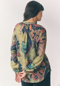 Lange mouwen bloes met bloemenprint in groen, rood en blauw. Geweven stof met een geplooide halslijn en elastische mouwboorden. De zoomlijn is gebogen.