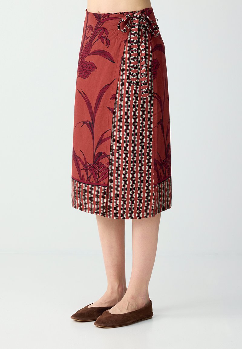 Jupe portefeuille longueur genou en rouge rouille avec motifs floraux et géométriques, nouée sur le côté, portée avec des chaussures plates marron.