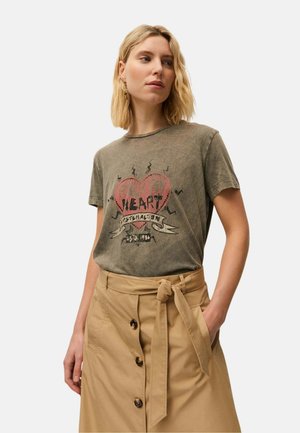 Mujer rubia con una camiseta gráfica de oliva desvanecida con diseño de corazón rosa metida en una falda beige abotonada con cinturón atado.