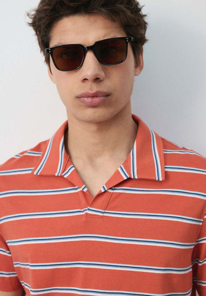 Jeune homme aux cheveux courts et bruns portant des lunettes de soleil foncées et un polo rayé orange sur un fond uni.
