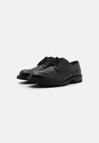 Chaussures en cuir noir avec des orteils arrondis, un laçage, et des semelles texturées. Présentent un design épuré et une construction minimaliste.