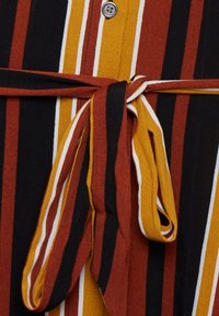 Tessuto a righe con linee verticali in nero, arancione, giallo e bianco. Il punto forte è un dettaglio con cintura legata in vita e chiusura con bottone.