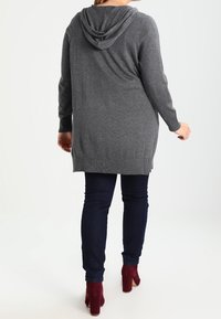 Czarnoszary długi rękaw z kapturem, z kieszenią z przodu i bocznymi rozcięciami, w połączeniu z ciemnymi dżinsami i burgundowymi botkami na obcasie. Miękki, teksturowany materiał.