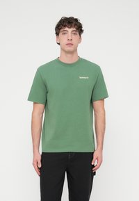 Πράσινο βαμβακερό μπλουζάκι με κοντά μανίκια, στρογγυλή λαιμόκοψη και μικρό χρυσό λογότυπο Timberland στο αριστερό στήθος. Απλή υφή, άνετη εφαρμογή.