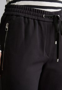 Zwarte joggingsbroek met een trekkoord in de taille, een ritszak aan de zijkant en een gladde, gestructureerde stof. Heeft een nauwsluitende pasvorm rond de heupen.