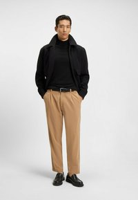 Giacca nera con colletto alto, dolcevita nera aderente, pantaloni beige e scarpe slip-on nere. Design semplice e moderno con linee pulite.
