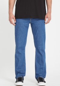 Pantalones vaqueros de mezclilla azul con corte recto, lavado medio, costuras prominentes, diseño estándar de cinco bolsillos, combinados con zapatillas negras.
