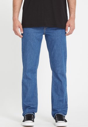Blue denim jeans with a straight cut, medium wash, prominent stitching, standard five-pocket design, worn with black sneakers.
