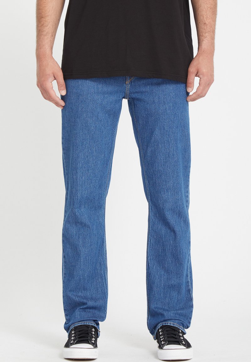 Pantalones vaqueros de mezclilla azul con corte recto, lavado medio, costuras prominentes, diseño estándar de cinco bolsillos, combinados con zapatillas negras.