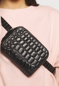 Borsa in pelle nera con effetto cocco, di forma rettangolare, con tracolla regolabile e chiusura con zip superiore, indossata a tracolla.