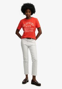 Czerwona bawełniana koszulka z białym napisem, zestawiona z białymi dopasowanymi jeansami i ciemnobrązowymi mokasynami. Modelka ma bujne czarne włosy.