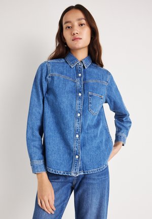 Chemise en denim à boutons de couleur bleu moyen, dotée d'un col classique, d'une poche poitrine, et de détails de couture subtils, associée à un jean assorti.