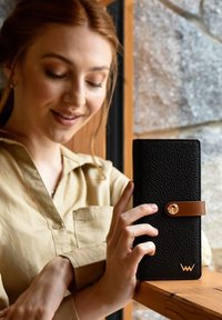 Black textured wallet with a brown leather strap and gold hardware accent, held by a person in a light shirt against a wooden surface.