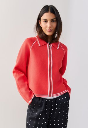 Vrouw draagt een rood jack met rits en witte bies en een zwarte broek met polkadots, staand met handen in de zakken tegen een effen achtergrond.