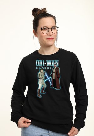 Kvinde med briller iført en sort sweatshirt med Obi-Wan Kenobi med lyssværd og tekst, stående mod en ensfarvet baggrund.