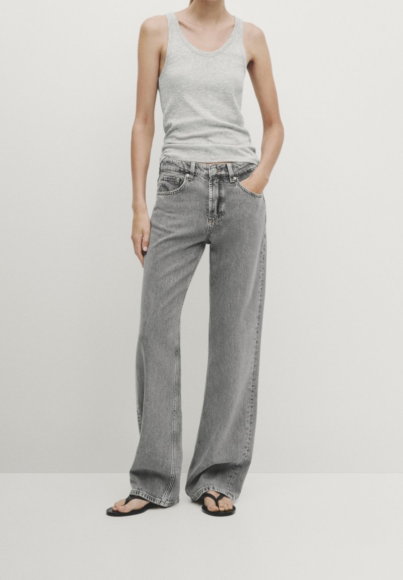 Débardeur gris clair associé à un jean en denim gris large, avec une taille haute et un design classique à cinq poches. Des tongs noires complètent le look.