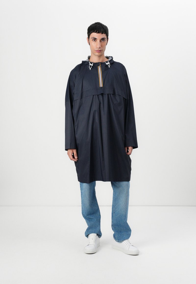 Marineblå vanntett poncho med hette, frontlynlås med gule og hvite striper, løs passform og lange ermer. Kombinert med lyseblå jeans.
