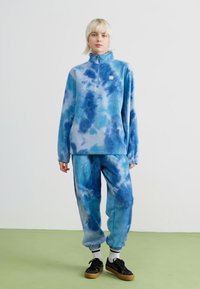 Blå tie-dye fleece-pullover med kvart glidelås og matchende joggebukser, med en avslappet passform og myk tekstur. Sorte sneakers og hvite sokker.