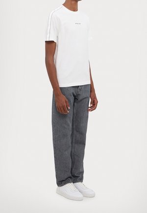 Man draagt een wit T-shirt met korte mouwen met zwarte strepen aan de zijkant, grijze rechte jeans en witte sneakers, staand tegen een effen achtergrond.