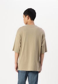 Beige oversized tričko vyrobené z bavlny. Má krátké rukávy, kulatý výstřih a rovný střih. Nosič má na sobě modré džíny.