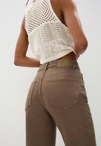 Braune hoch taillierte Hose mit glatter Textur und zwei Gesäßtaschen. Mit einem hellen Etikett an der Taille und einem gestrickten cremefarbenen Oberteil mit offener Webart.