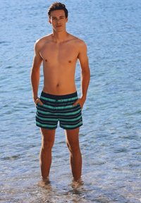 Shorts de bain pour hommes avec des rayures teal et marine, taille à cordon, longueur mi-cuisse, portés dans une eau peu profonde avec un arrière-plan bleu clair.