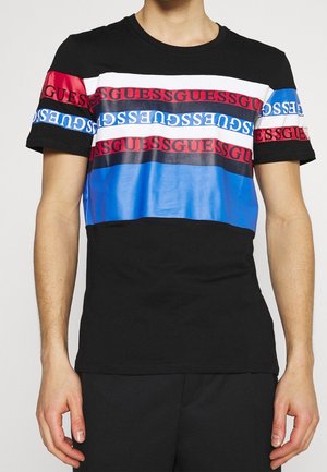 Persoon draagt een zwart T-shirt met horizontale rode, witte en blauwe strepen en herhaalde tekst "GUESS" over de borst en mouwen.