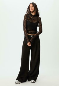 Sort blonde lace-top med lange ærmer, der har udskæringsdetaljer og lagdelt design, parret med løstsiddende sorte bukser og sølvfarvede sneakers.
