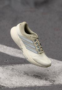 Μπεζ ελαφρύ παπούτσι για τρέξιμο με αναπνεύσιμο επάνω μέρος, που διαθέτει τρεις μπλε ρίγες, υφή σόλας και κομψό, αεροδυναμικό σχεδιασμό.