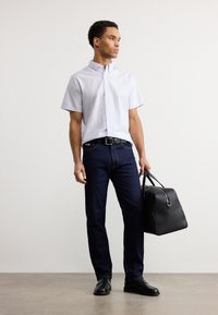 Ανοιχτό μπλε πουκάμισο με κοντά μανίκια και κουμπιά, σκούρο τζιν τζιν, μαύρη ζώνη και μαύρα παπούτσια. Κρατάει μια στιβαρή μαύρη τσάντα με κορυφαίες λαβές.