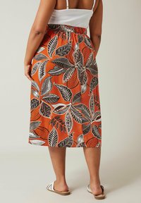 Falda midi con un fondo naranja y patrones de hojas en blanco y negro, cintura elástica y corte relajado. Llevada con sandalias planas.