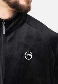 Zwarte zip-up jas van zachte velvet. Beschikt over een hoge kraag en een wit geborduurd logo op de borst. Gladde textuur aan de hele jas.