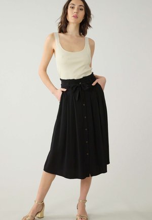 Jupe midi noire avec des plis, des boutons sur le devant et une taille nouée. Portée avec un débardeur beige et des sandales à talons blocs dorées.