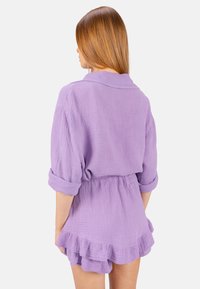 Lila strukturiertes Stoffoutfit, das ein Hemd mit hochgekrempelten Ärmeln und einen Rock mit Rüsche enthält. Der elastische Bund sorgt für Komfort und perfekten Sitz.