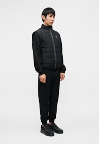 Μαύρο γιλέκο με καπιτονέ ύφασμα χωρίς μανίκια και ψηλό γιακά, σε συνδυασμό με μαύρο μακρυμάνικο πουκάμισο και μαύρο παντελόνι στενής γραμμής· ομοιόχρωμο σε όλο το σύνολο.