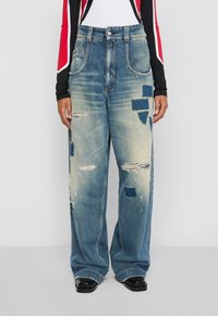 Široké vyblednuté modré džínsy s opotrebovanými škvrnami a detailmi, na ktorých sa nachádza osoba v dlhom rukávovom čiernom a červenom topu a čiernych topánkach.