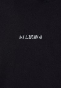 Černé bavlněné tričko s žebrovanou texturou a bílým vyšívaným nápisem „HAN KJØBENHAVN“ uprostřed na hrudi.