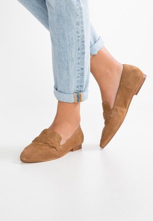 Persoon die lichtblauwe opgerolde jeans en bruine suède loafers draagt, staand op een witte ondergrond met één voet lichtjes opgeheven.