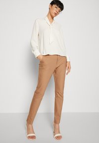 Cremefarbige Bluse mit Knoten am Ausschnitt, kombiniert mit taillierten beigefarbenen Hosen und weißen Sandalen mit Blockabsatz. Einfaches und elegantes Design.