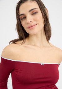 Burgundské off-the-shoulder tričko s ribetkovejšou textúrou, svetlomodrý lem pozdĺž výstrihu a malá dekoratívna mašľa v strede.